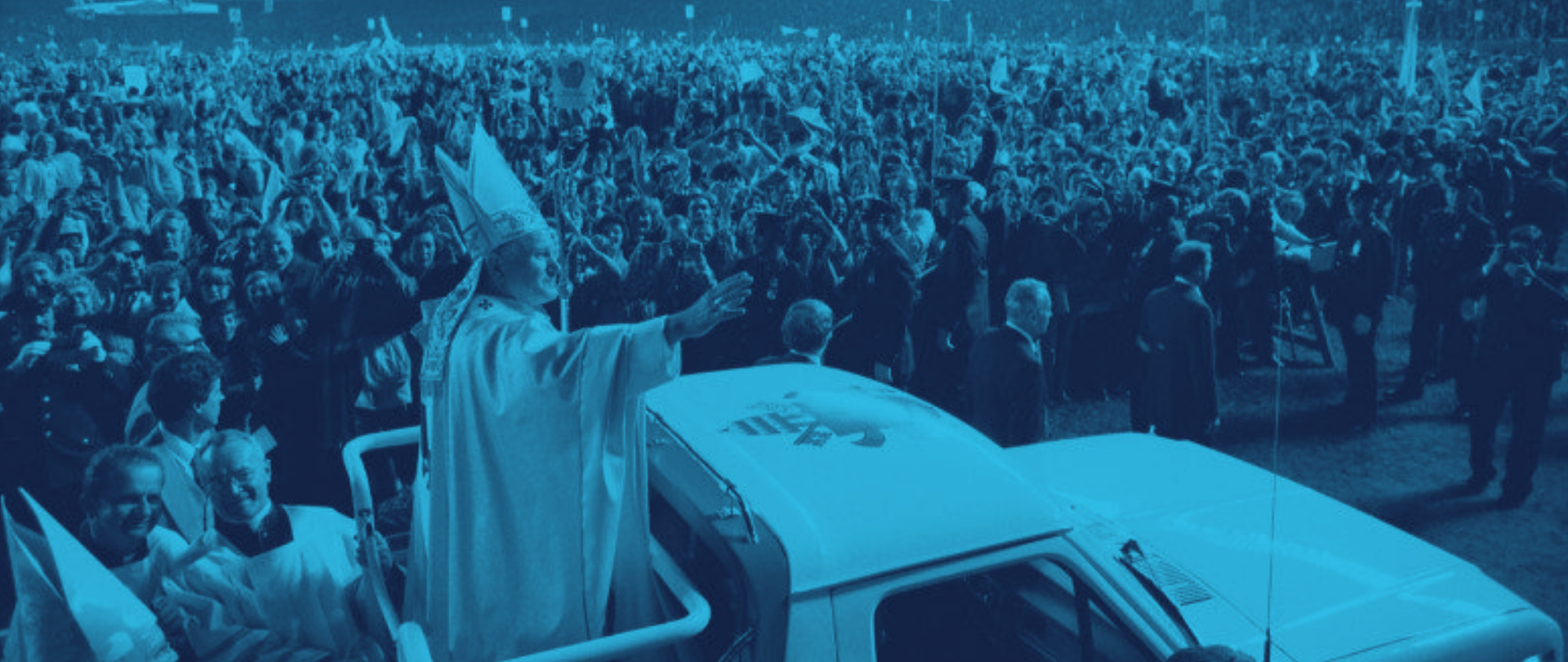A foto apresenta o Papa João Paulo II num papamóvel aberto. O homem está vestido de uma casula festiva e uma mitra. Com a mão direita erguida na altura dos ombros, cumprimenta a multidão que o recebe.