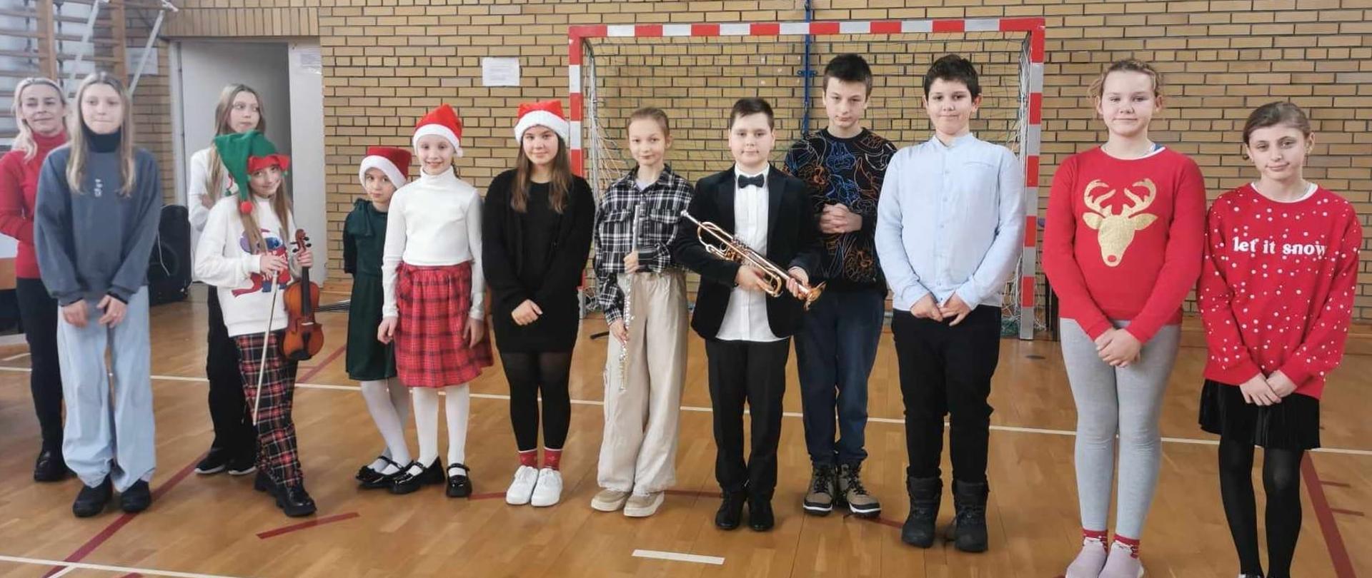Zdjęcie grupowe zrobione na sali gimnastycznej. Uczeń na środku trzyma trąbkę, dziewczyna po lewej stronie trzyma skrzypce.