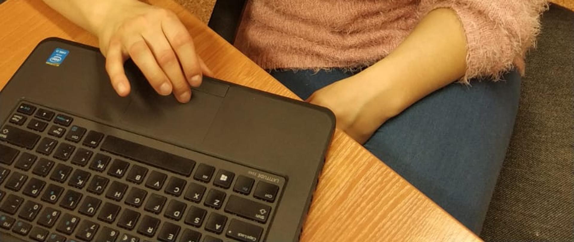 Przedstawia kobietę w różowym swetrze i w niebieskich jeansach, siedzącą przy laptopie, prowadzącą zajęcia.