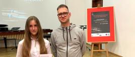 Zdjęcie przedstawia nauczyciela i uczennicę z dyplomem w sali koncertowej w Stargardzie podczas konkursu perkusyjnego. W tle telebin i sztaluga z plakatem konkursu.