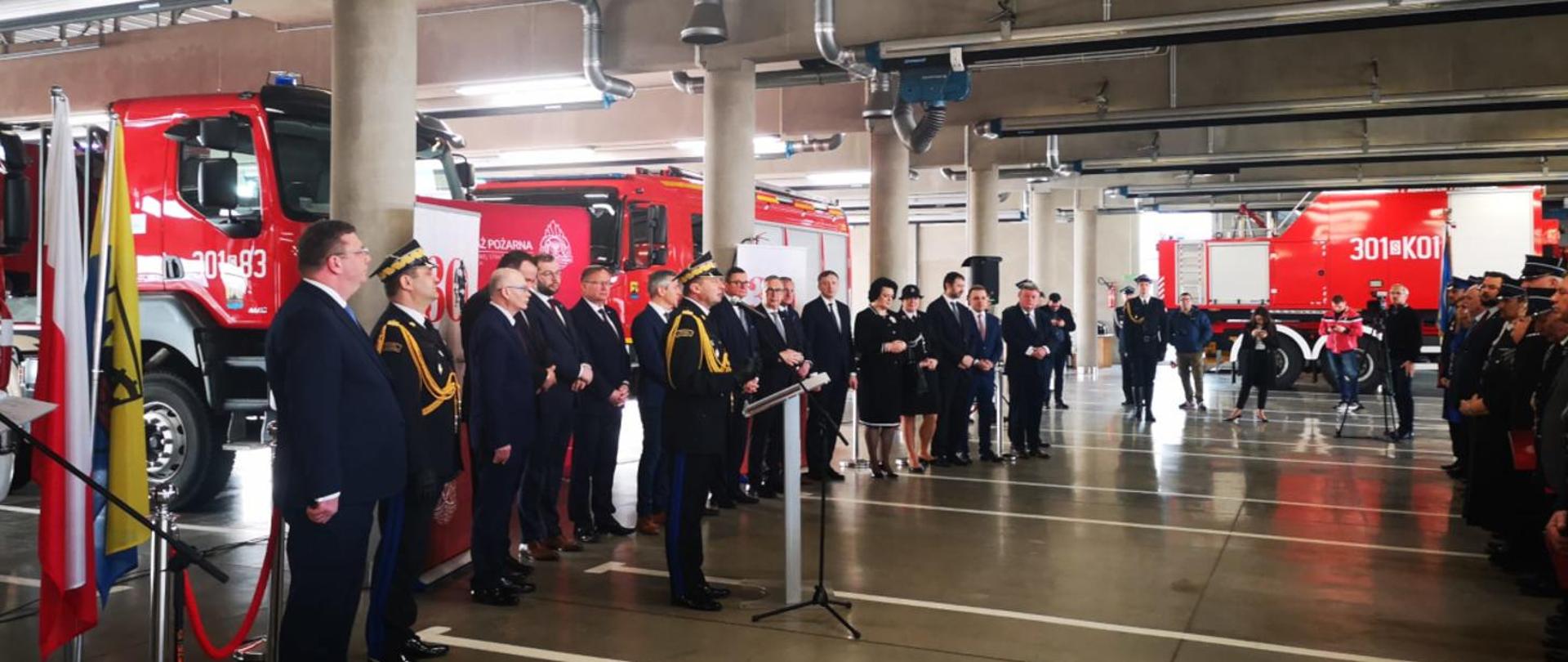 Zdjęcie zrobione w pomieszczeniu garażowym strażnicy na tle samochodów pożarniczych. Na pierwszym planie strażacy w umundurowaniu galowym oraz zaproszeni na uroczystość goście.