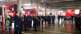 Zdjęcie zrobione w pomieszczeniu garażowym strażnicy na tle samochodów pożarniczych. Na pierwszym planie strażacy w umundurowaniu galowym oraz zaproszeni na uroczystość goście.