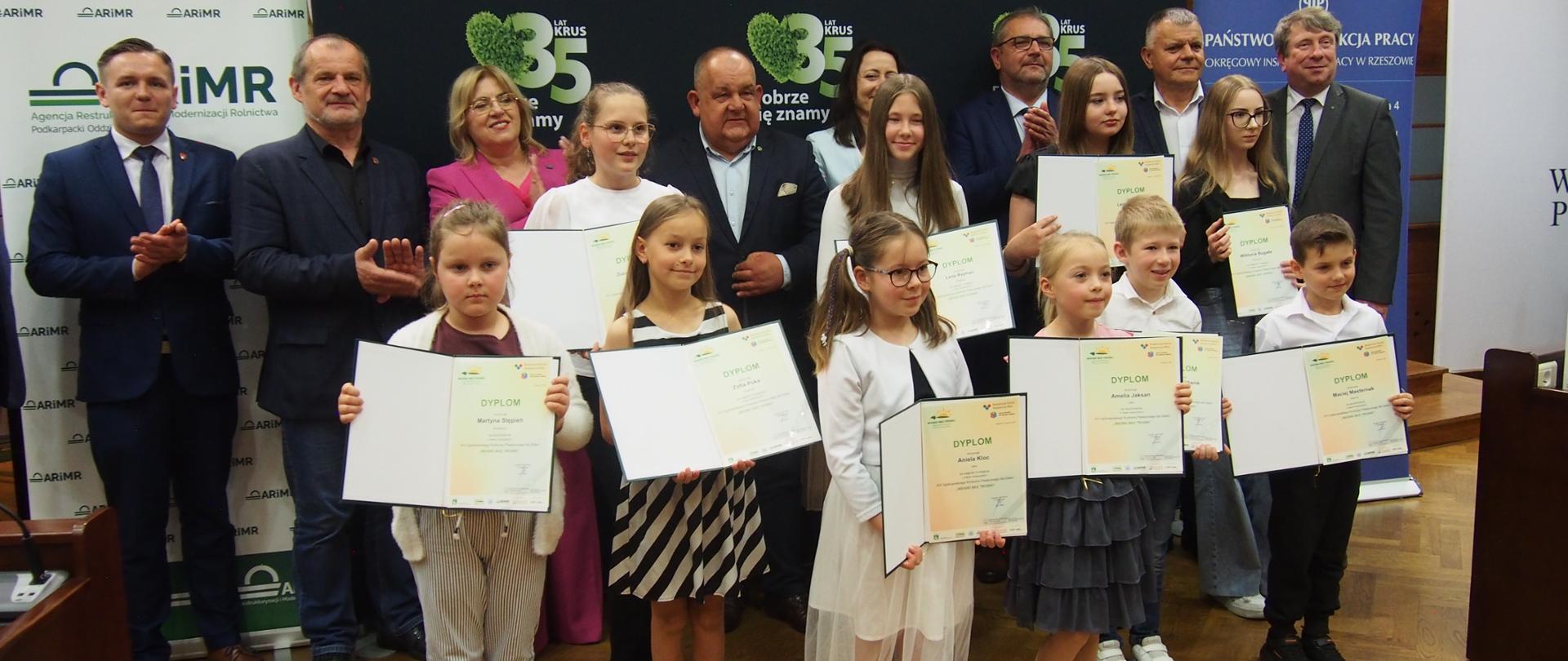 Grupa osób – laureaci konkursu wraz z przedstawicielami instytucji na tle roll-upów KRUS, PIR oraz ARiMR pozują do zdjęcia. Dzieci – laureaci konkursów trzymają w rękach dyplomy