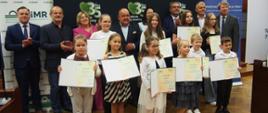 Grupa osób – laureaci konkursu wraz z przedstawicielami instytucji na tle roll-upów KRUS, PIR oraz ARiMR pozują do zdjęcia. Dzieci – laureaci konkursów trzymają w rękach dyplomy