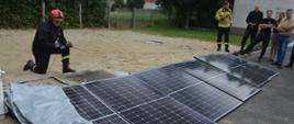 Warsztaty z zakresu fotowoltaiki. Plac Komendy Powiatowej PSP w Rawiczu, na którym odbywa się część praktyczna. Prowadzący prezentuje zwarcie. Przed nim ułożone są panele fotowoltaiczne. Strażak na głowie ma czerwony hełm. Z prawej strony stoją uczestnicy warsztatów.