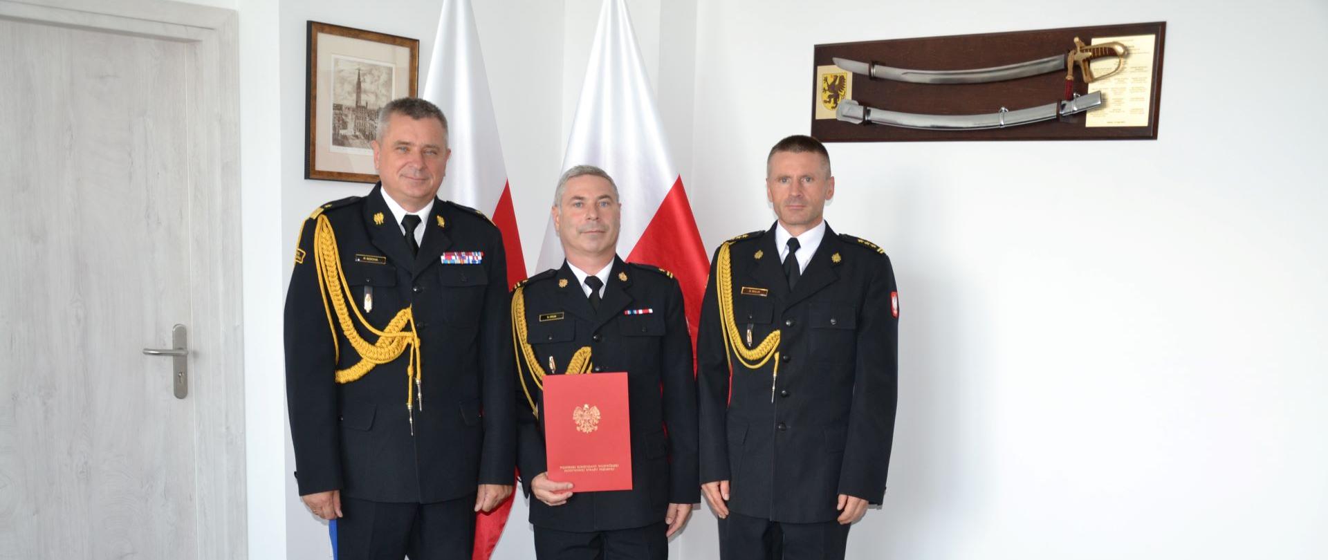 Pomorski komendant wojewódzki Państwowej Straży Pożarnej stoi wraz ze strażakami jeden z nich trzyma czerwoną teczkę za nimi stoją flagi Polski.