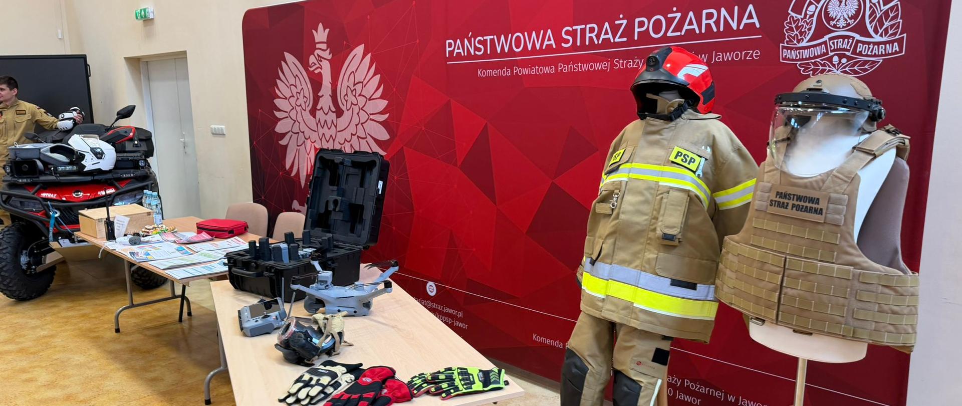 Na tle czerwonego baneru służb pożarniczych ustawiono dwa manekiny prezentujące różne elementy wyposażenia strażaków — jeden w pełnym ubraniu ochronnym z hełmem, drugi w kamizelce taktycznej z osłoną twarzy. Na stołach przed nimi rozłożono sprzęt ratowniczy, w tym rękawice, narzędzia i akcesoria techniczne. Po lewej stronie stoi funkcjonariusz w beżowym mundurze obok czerwonego pojazdu terenowego wyposażonego w dodatkowy sprzęt. Scena przedstawia ekspozycję wyposażenia straży pożarnej.