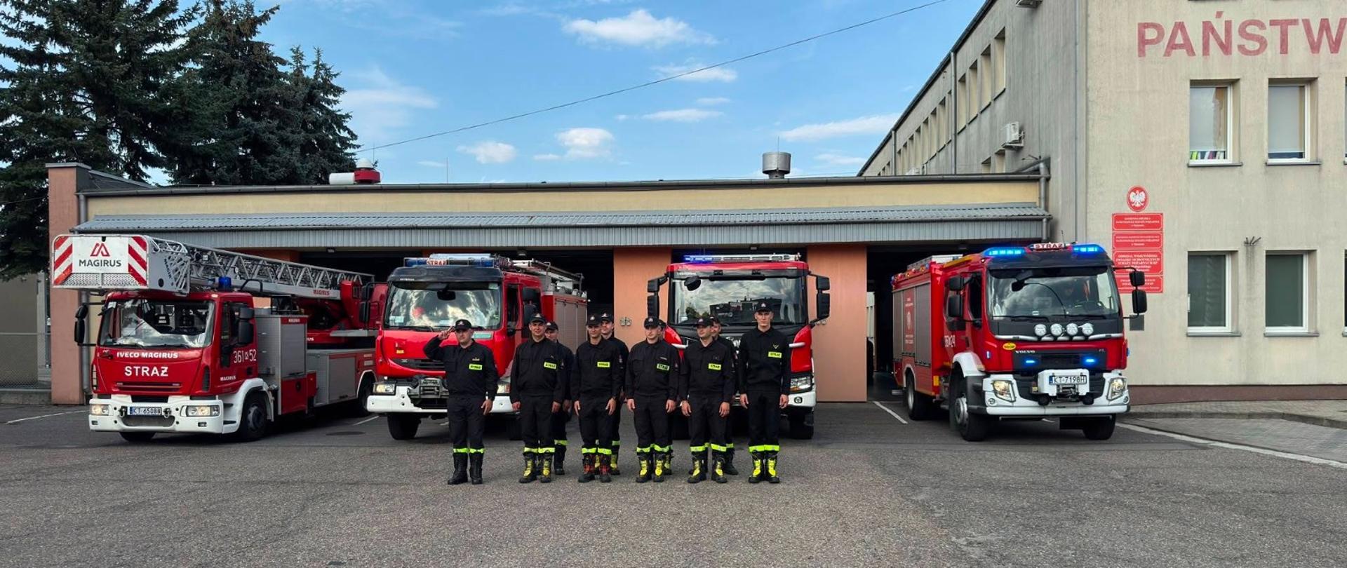 Przed samochodami strażackimi stoją w dwuszeregu strażacy ubrani w czarne ubrania koszarowe na głowach mają czapki z daszkiem. W tle budynek remizy i plac