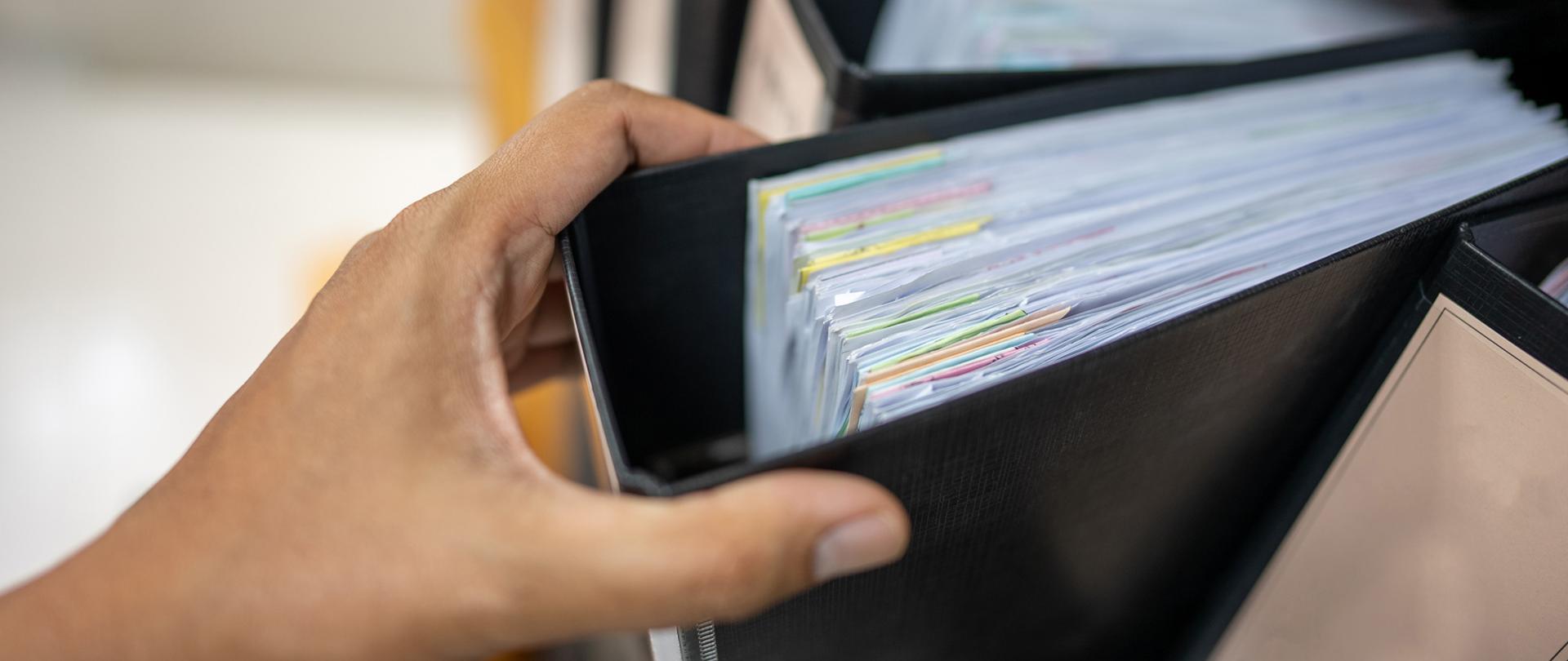 Dłoń sięga po jeden z szeregu segregatorów z dokumentami.