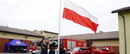Trzech funkcjonariuszy Państwowej Straży Pożarnej w galowych mundurach uczestniczy w ceremonii podniesienia biało-czerwonej flagi. W tle widoczne są czerwone wozy strażackie, namiot Komendy Powiatowej Państwowej Straży Pożarnej oraz sprzęt ratowniczy ustawiony na placu jednostki.