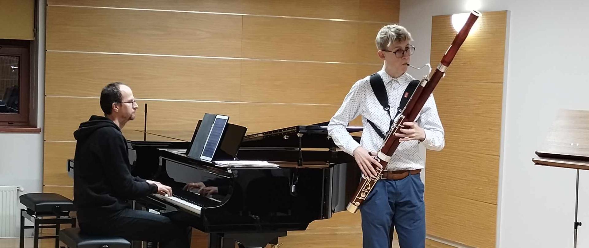 Na scenie sali kameralnej uczeń gra na fagocie. Z prawej strony pusty pulpit. Z lewej strony dwa fortepiany, przy jednym z nich gra akompaniator spoglądający na nuty. W tle z lewej strony widoczna część okna a pod nim kaloryfer.