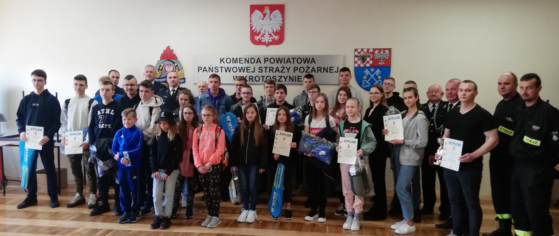 Zdjęcie grupowe na świetlicy KP PSP. Na zdjęciu uczestnicy konkursy czyli młodzież szkolna oraz organizatorzy. Dzieci trzymają w rękach nagrody oraz dyplomy. 