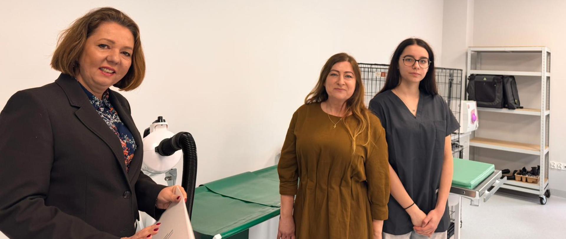 Do zdjęcia pozują 3 osoby, w tym wiceminister I. Ziętka. W tle widać kozetki lekarskie, klatki weterynaryjne oraz półki z różnymi przedmiotami. Wiceminister Ziętka w dłoni trzyma teczkę z logiem Ministerstwa Edukacji Narodowej.