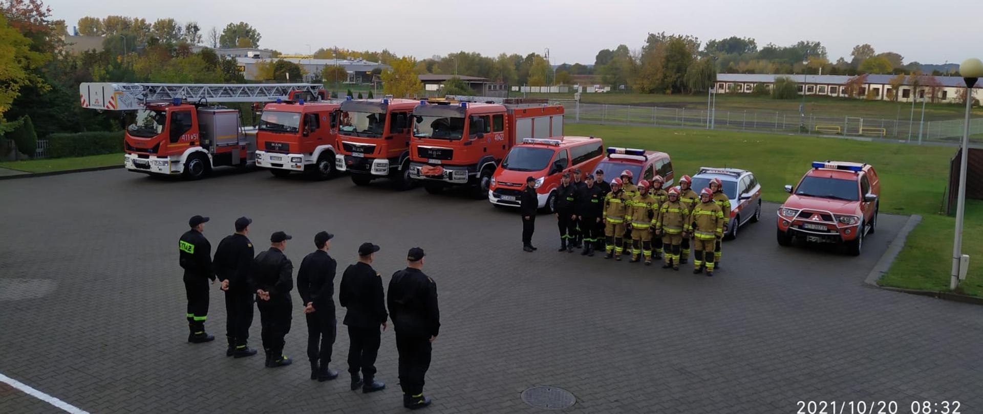 Zbiórka strażaków przed samochodami.