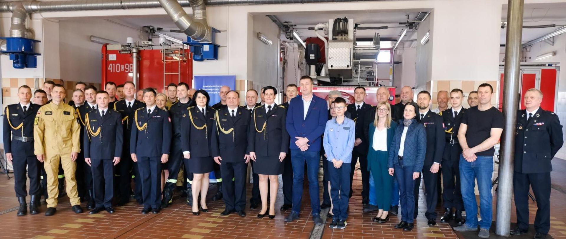 Zdjęcie grupowe uczestników uroczystości w garażu KP PSP Chojnice na tle samochodów pożarniczych. 
