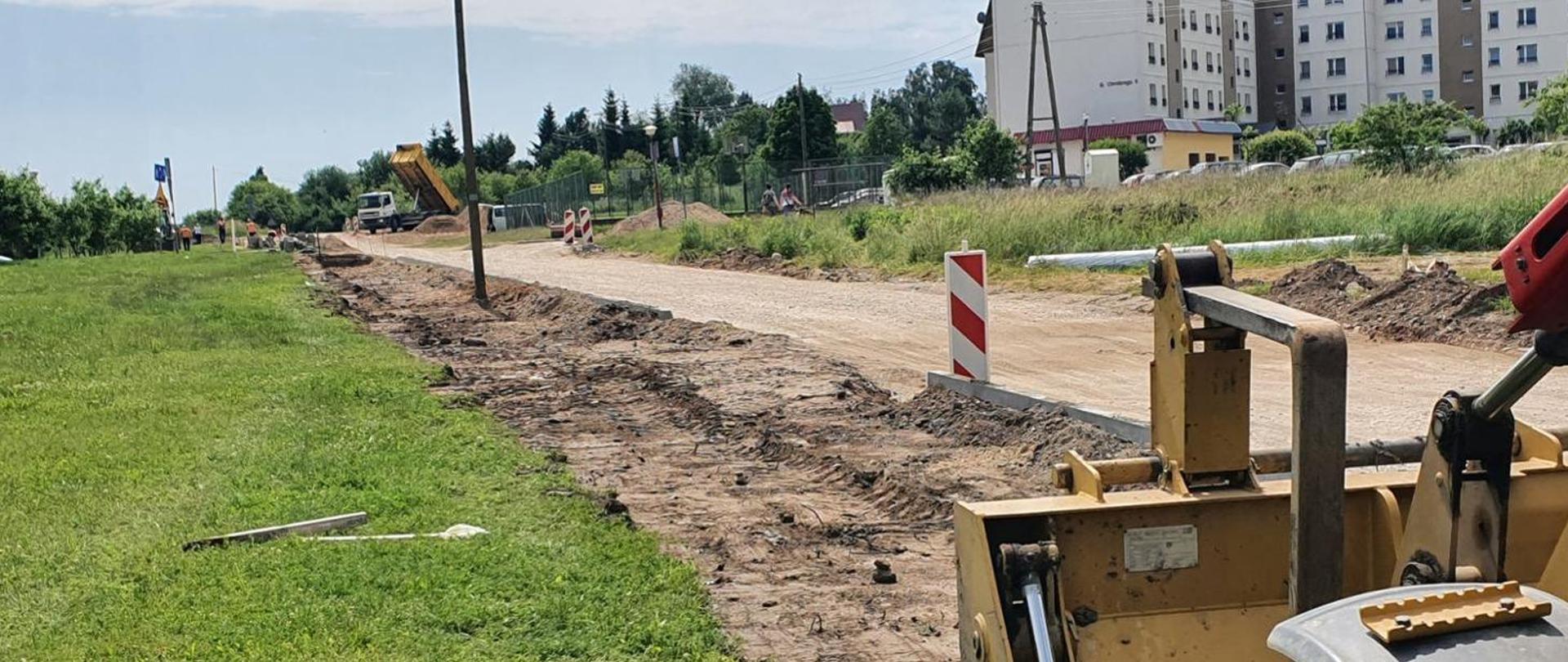Rozkopana droga. Na pierwszym planie widoczna jest łopata koparki. W tle osiedle bloków