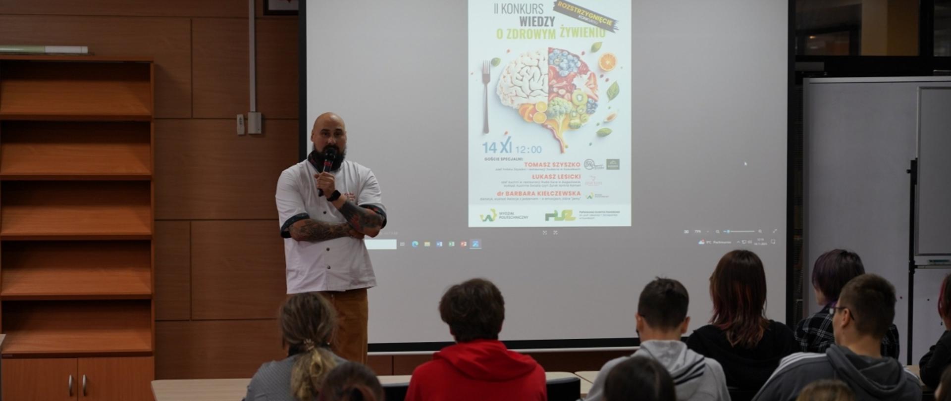 Prelegent z mikrofonem, stojący w białym kitlu kucharza, prowadzi prezentację dla grupy młodzieży siedzącej w auli lub sali lekcyjnej. W tle na dużym ekranie projektor wyświetla plakat II Konkursu Wiedzy o Zdrowym Żywieniu.