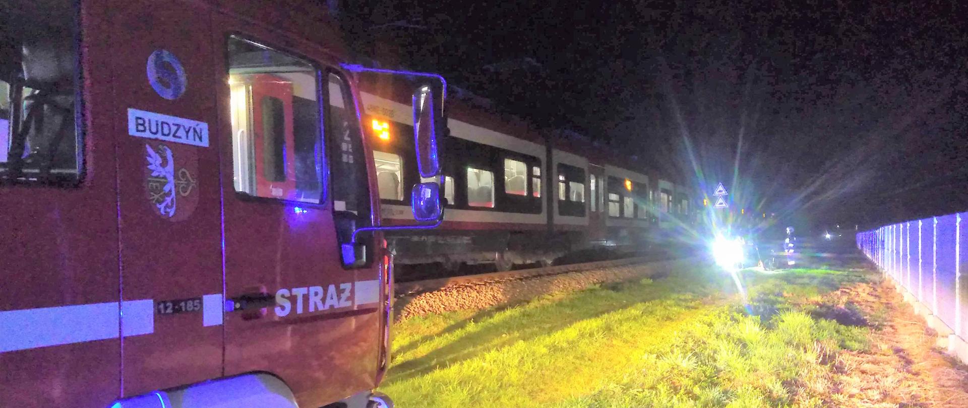 Zdjęcie przedstawia pociąg i samochód strażacki, stojące w polu z powodu śmiertelnego potracenia mężczyzny.
W tle pole i płot.
