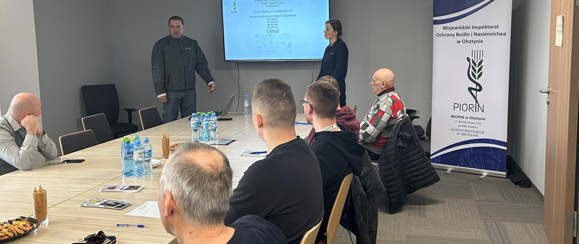 grupa ludzi siedzących przy stole. Na środku pomieszczenia na ścianie, wisi ekran, na którym wyświetlana jest prezentacja. Kobieta i mężczyzna stoją przed ekranem i prowadzą prezentacje. W tle widoczny jest roll-up z napisem „PIORiN” oraz logo organizacji