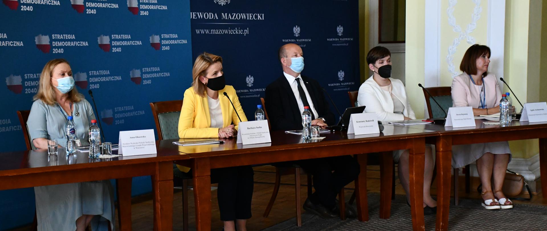 Na zdjęciu znajduje się pięć elegancko ubranych osób - cztery kobiety i mężczyzna. Osoby te siedzą za stołem prezydialnym, na tle ścianek wizerunkowych Wojewody Mazowieckiego oraz projektu "Strategia demograficzna 2040". 