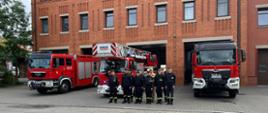 Trzy wozy strażackie, w tym drabina mechaniczna, stoją przed budynkiem strażnicy z czerwonej cegły; przed nimi ustawieni są strażacy w czarnych i khaki mundurach w szyku.