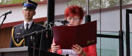 kobieta odczytuje list gratulacyjny do mikrofonu stojąc przy mównicy, w tle stoi strażak ubrany w umundurowanie wyjściowe z czapka rogatywką na głowie