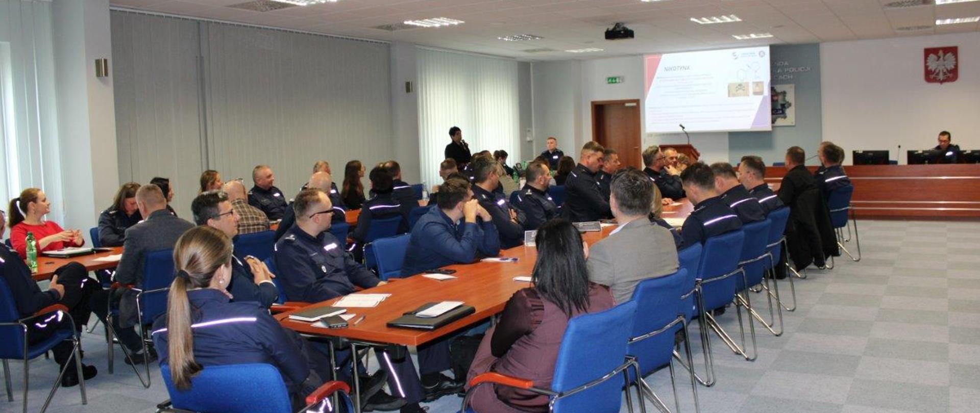 Na pierwszym planie uczestnicy konferencji - policjanci woj. świętokrzyskiego ubrani w większości w mundury policyjne siedzący przy stołach i słuchający wykładu. W tle przedstawicielka WSSE w Kielcach stojąca i prezentująca wykład wyświetlony na ekranie.