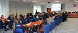 Na pierwszym planie uczestnicy konferencji – policjanci woj. świętokrzyskiego ubrani w większości w mundury policyjne siedzący przy stołach i słuchający wykładu. W tle przedstawicielka WSSE w Kielcach stojąca i prezentująca wykład wyświetlony na ekranie.