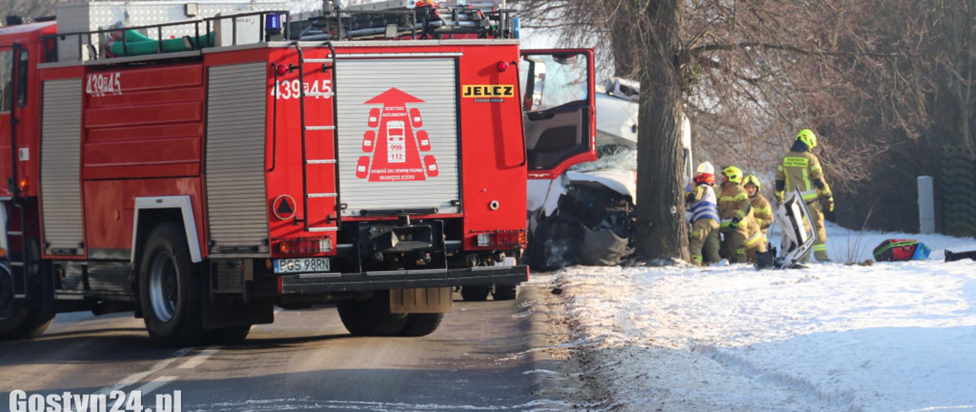 Na pierwszym planie stoi wóz strażacki. W tle są strażacy, którzy prowadzą działania przy uszkodzonym pojeździe.