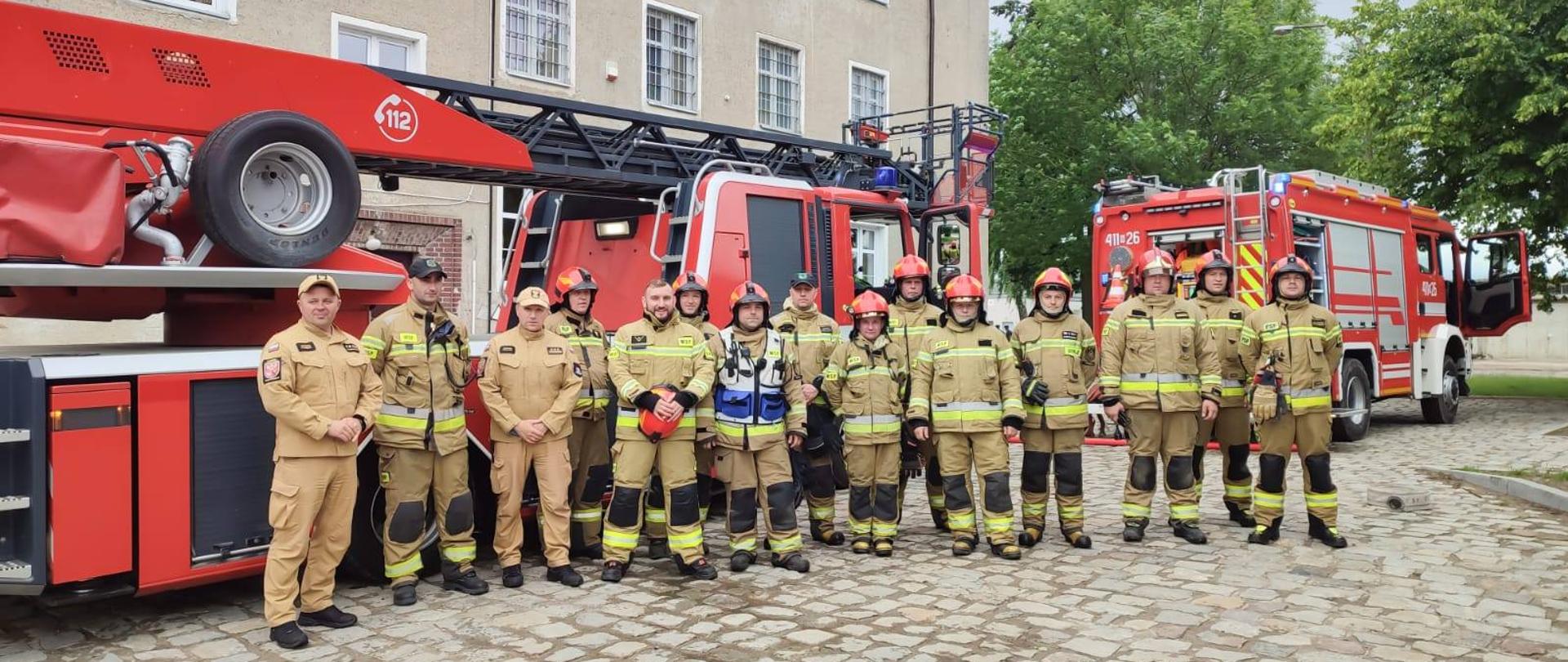 Grupa kilkunastu strażaków w mundurach bojowych stoi obok siebie na tle samochodów strażackich.