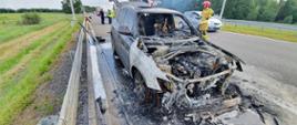 Pożar auta na S5. Jezdnia trasy S5 w kierunku Poznania. Na pasie awaryjnym stoi wrak spalonego samochodu osobowego. Obok niego strażak, a za nim policjanci i osoby cywilne. Lewym pasem drogi poruszają się pojazdy. W tle łąka oraz las.