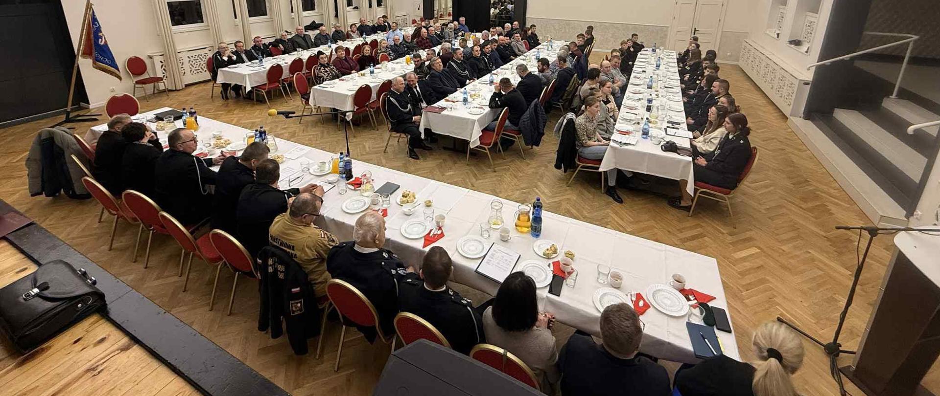 Strażacy w mundurach wyjściowych oraz cywile siedzący przy 5 stołach na sali bankietowej.