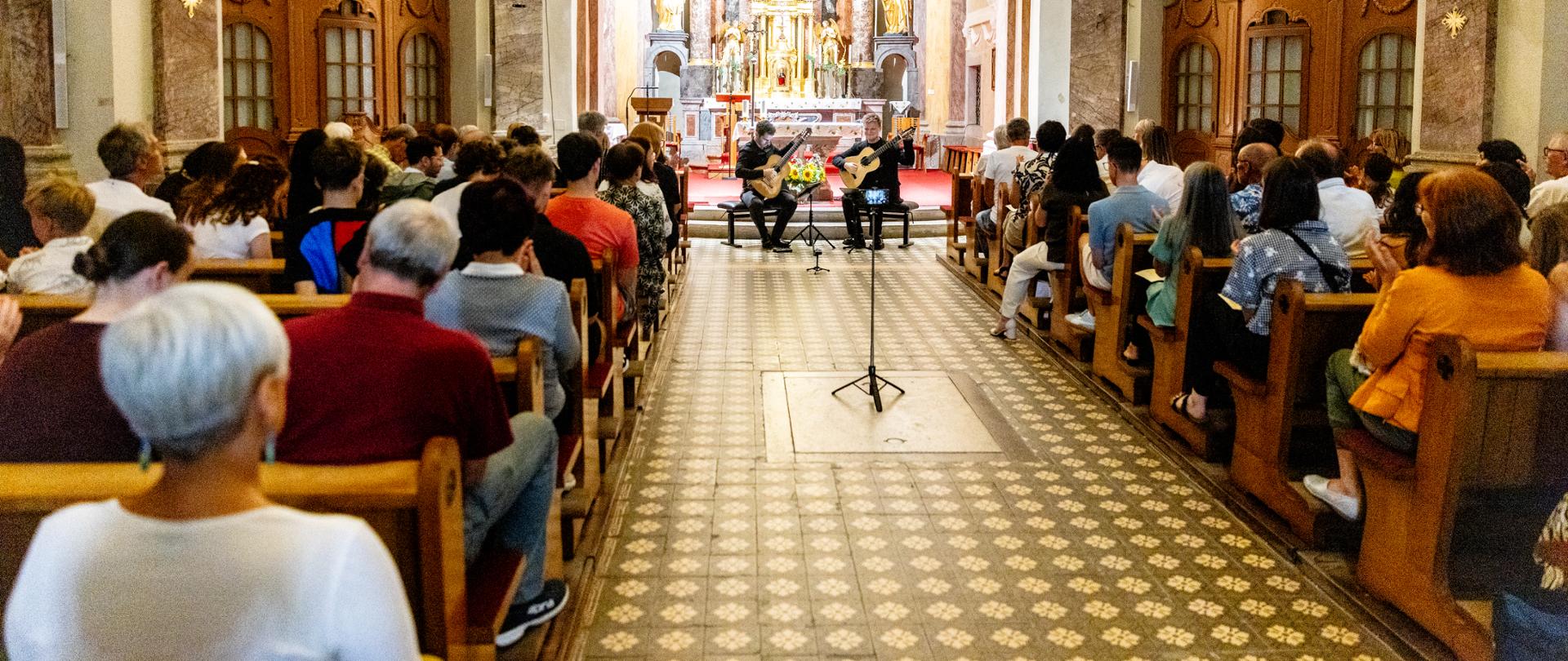 Notranjost baročne cerkve sv. Štefana v Postojni je polna občinstva. Pred glavnim oltarjem sedita kitarista Mak Grgić in Mateusz Kowalski ter izvajata koncert. Pogled z zadnjega dela cerkve poudarja številčno občinstvo in zbrano vzdušje.