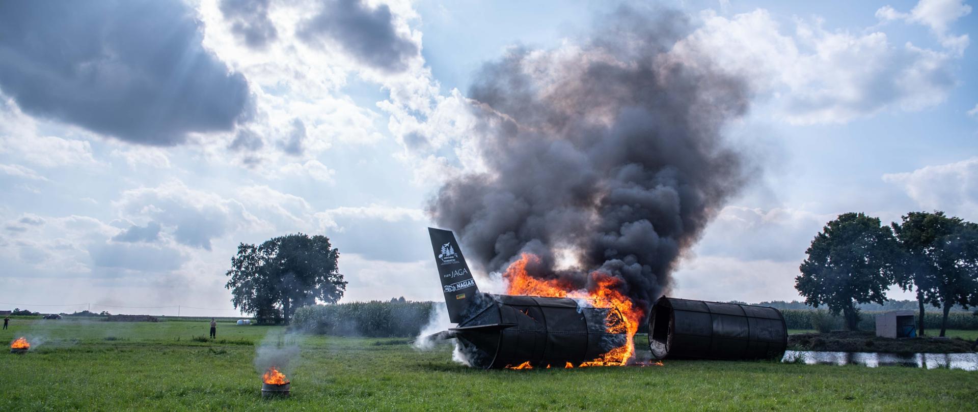 Wrak samolotu, który płonie. Samolot wykonany z metalowych silosów na potrzeby pozoracji. Samolot znajduje się nad zbiornikiem wodnym. Widcznie duże płomienie i gęsty dym.