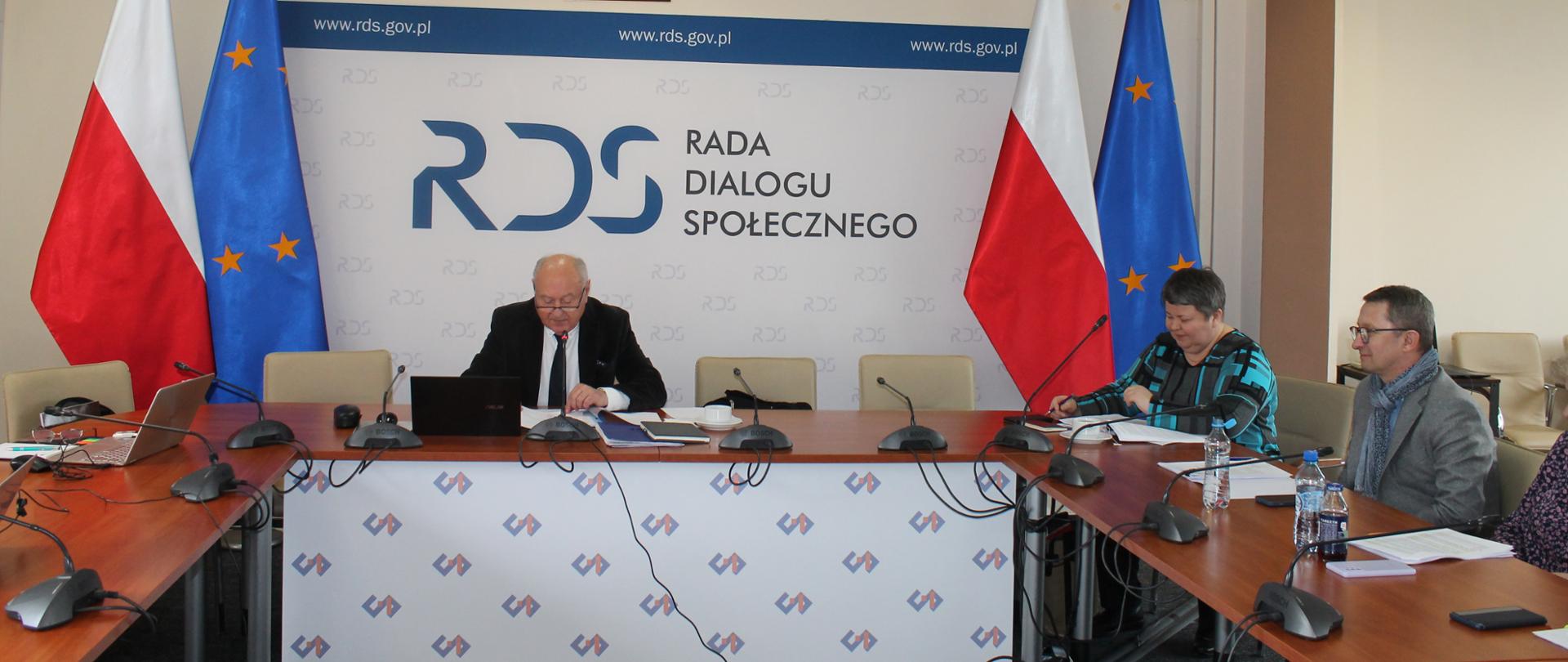 Na zdjęciu widnieje stół prezydialny za którym po środku Przewodniczący Zespołu ds. rozwoju dialogu społecznego a po Jego lewej przedstawicielka Związku Rzemiosła Polskiego i dalej Dyrektor Departamentu Dialogu i Partnerstwa Społecznego w MRPiPS. Po obu stronach stołu widnieją dwie flagi: Polski i UE. W tle siedzących znajduje się baner z napisem Rada Dialogu Społecznego a nad nim wisi godło Polski.
