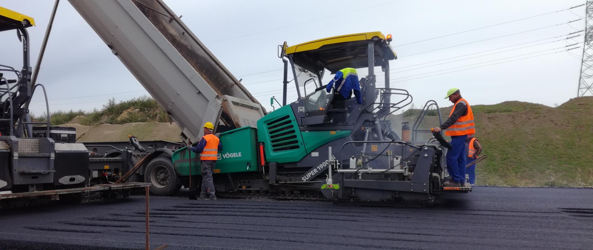 Zdjęcie przedstawia układanie masy bitumicznej z bliska. Na zdjęciu widoczne są rozściełacze czyli maszyny do układania masy i pracujący pracownicy. Widoczne są warstwy drogi podbudowa, i warstwa wiążąca.