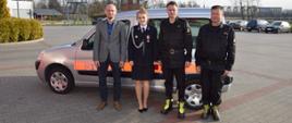 Zdjęcie w formacie panoramicznym. Zdjęcie na tle przekazywanego samochodu SLKw Citroen Berlingo (koloru srebrnego z czerwonym napisem straż). Na zdjęciu przed pojazdem stoją od lewej Zastępca Wójta Gminy Ryńsk Pan Łukasz Gapiński, Pani Prezes Zarządu OSP Zieleń dh Judyta Majewska-Nowek, Komendant Powiatowy st. kpt. Robert Stasiak oraz Zastępca Komendanta Powiatowego st.bryg. Cezary Maćczak.