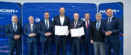 Zdjęcie zbiorowe, w rzędzie stoi ośmiu mężczyzn w garniturach, trzeci od lewej minister Kulasek, dwóch pośrodku trzyma otwarte teczki z dokumentami, za nimi granatowa ścianka z napisem NCBR.