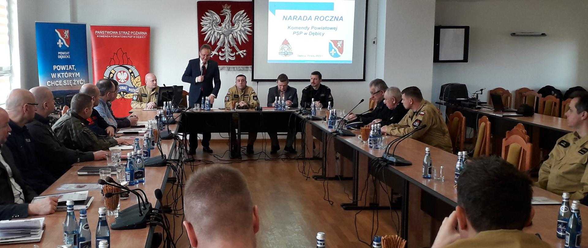 Straży oraz zaproszeni gości znajdują się w sali narad. Siedzą za ułożonymi w prostokąt stołami. Przemawia komendant powiatowy Państwowej Straży Pożarnej. Na ekranie wyświetlana jest prezentacja.