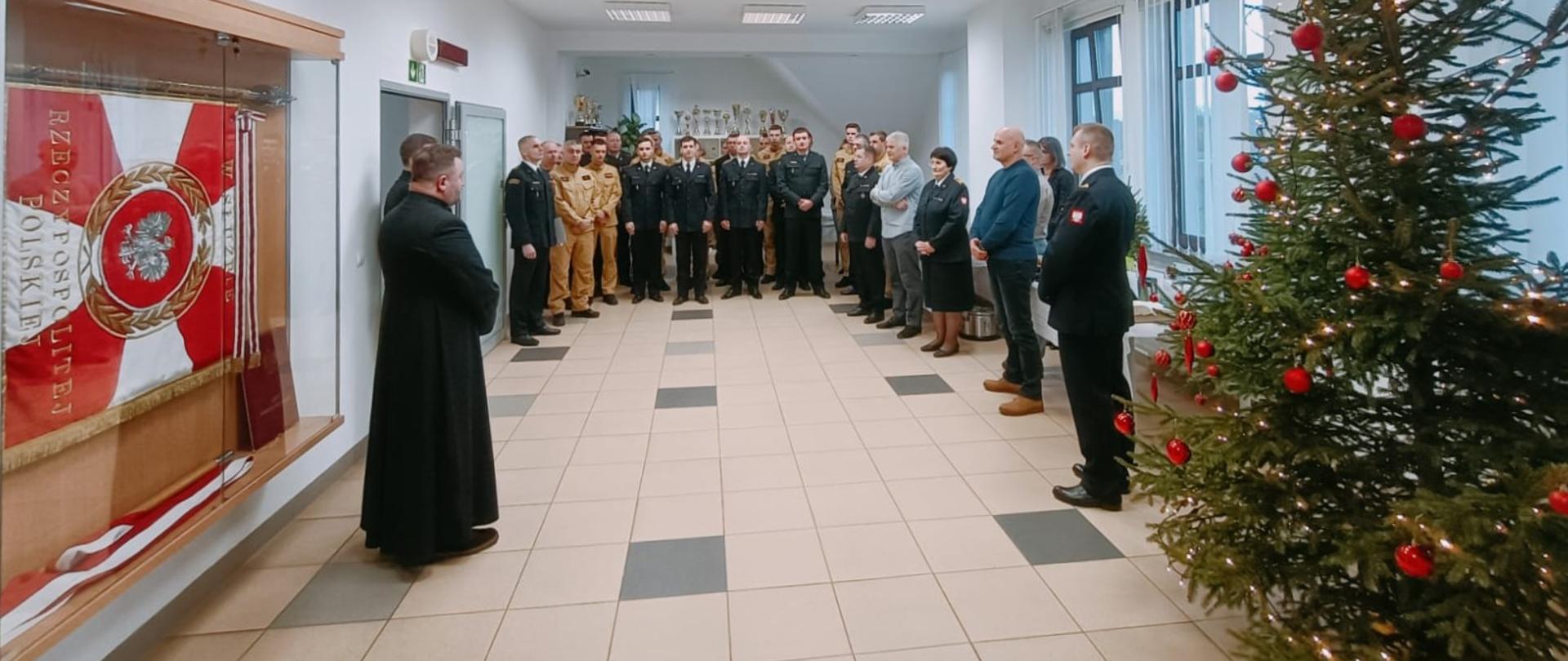 Zdjęcie przedstawia uczestników strażackiego spotkania opłatkowego (funkcjonariusze, pracownicy cywilni oraz zaproszeni goście) zgromadzonych w świetlicy Komendy Powiatowej Państwowej Straży Pożarnej w Gołdapi podczas składania wszystkim obecnym i Ich rodzinom życzeń z okazji Świąt Bożego Narodzenia oraz podziękowań za współpracę i wsparcie w mijającym roku przez Komendanta Powiatowego Państwowej Straży Pożarnej w Gołdapi st. bryg. Wojciecha Szczepanika