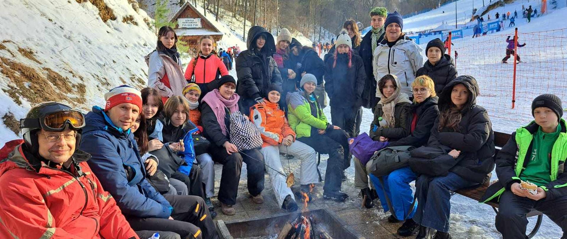 Fotografia. W Kazimierzu Dolnym u podnóża ośnieżonego stoku siedzą lub stoją uczniowie i nauczyciele: Tomasz Krasowski, Teresa Skrzypiec-Rejzner, Andrzej Mazuś. Znajdują się w półokręgu. W środku płonie ognisko. Większość z nich ubrana jest w stroje narciarskie. W tle: stok, narciarze, drzewa, ogrodzenie z siatki wyznaczające trasę narciarską, itd.