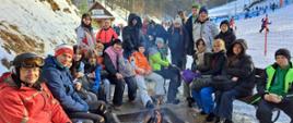 Fotografia. W Kazimierzu Dolnym u podnóża ośnieżonego stoku siedzą lub stoją uczniowie i nauczyciele: Tomasz Krasowski, Teresa Skrzypiec-Rejzner, Andrzej Mazuś. Znajdują się w półokręgu. W środku płonie ognisko. Większość z nich ubrana jest w stroje narciarskie. W tle: stok, narciarze, drzewa, ogrodzenie z siatki wyznaczające trasę narciarską, itd.