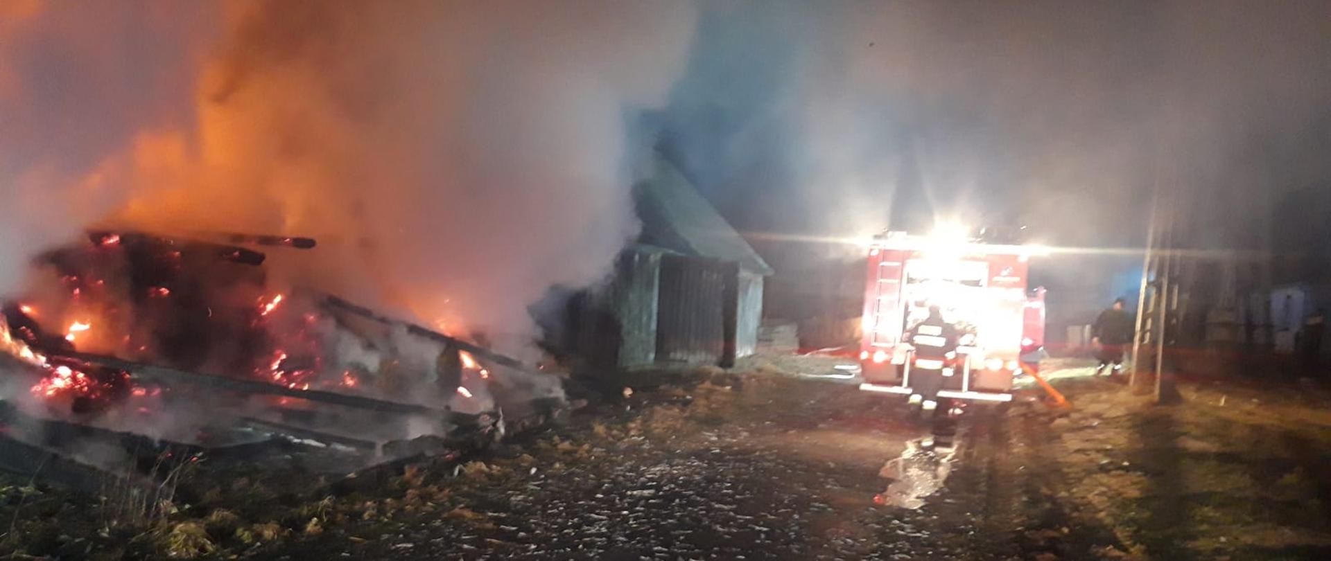 Pożar drewnianej stodoły. Widać płomienie i dym. Na drodze dojazdowej po prawej stronie stoi samochód strażacki.