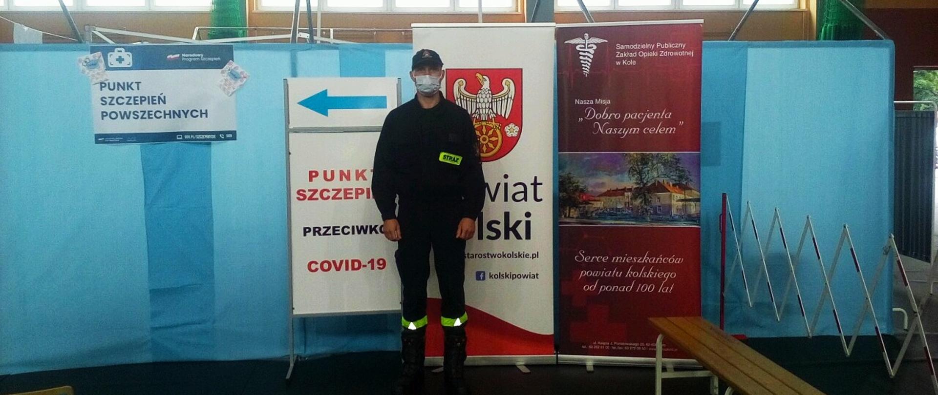 Zdjęcie przedstawia poprawnie umundurowanego strażaka na hali sportowej, gdzie zorganizowany jest punkt szczepień.