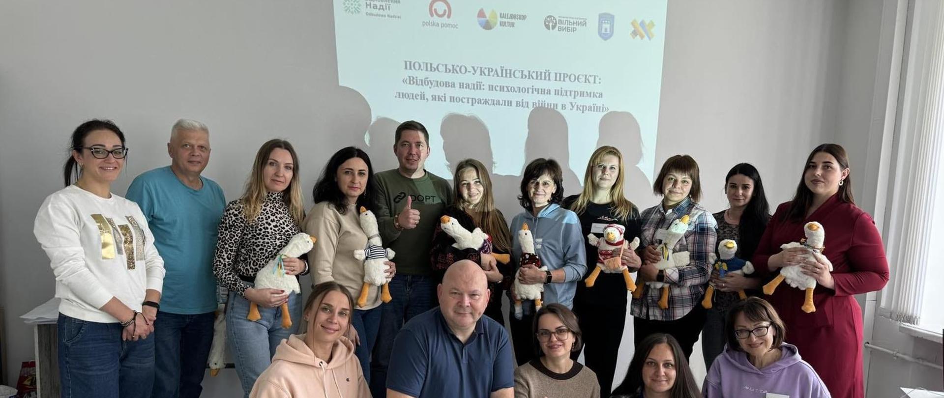 Grupa kobiet i mężczyzn pozuje do zdjęcia na tle slajdu, na którym wyświetlone jest logo Polskiej pomocy