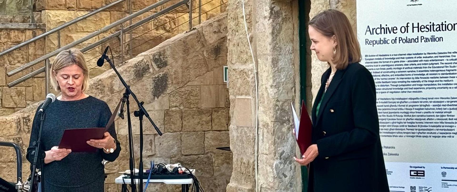 Pani Bożena Żelazowska, Sekretarz Stanu w Ministerstwie Kultury, czyta przemówienie podczas otwarcia Polskiego Pawilonu na Malta Biennale 2026. Przy drugim mikrofonie stoi profilowo druga kobieta, oczekująca na swoją kolej.