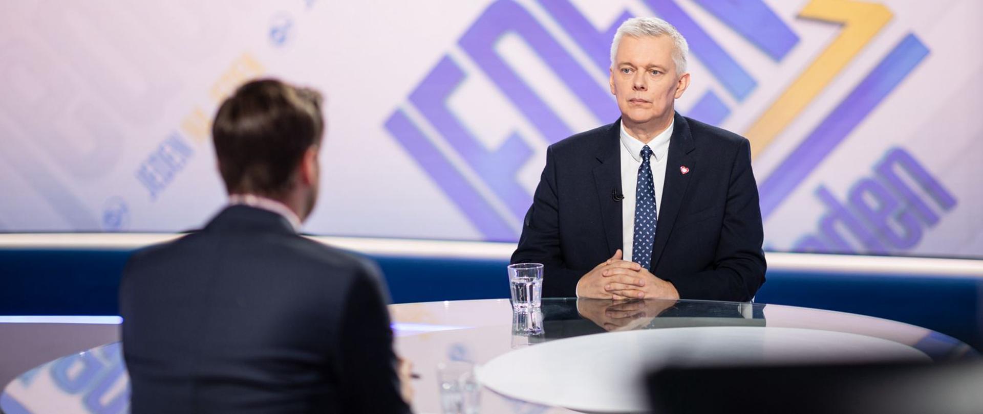 Minister Tomasz Siemoniak w garniturze siedzi przy stole podczas telewizyjnego wywiadu. Naprzeciwko niego, tyłem do obiektywu, siedzi prowadzący. W tle widoczne jest kolorowe studio z dużym napisem na ekranie.