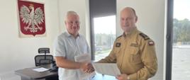Na zdjęciu widoczni są dwaj mężczyźni siedzący przy ciemnym stole biurowym i podpisujący dokumenty: Osoba po prawej stronię: Mężczyzna w piaskowym/beżowym mundurze służbowym Państwowej Straży Pożarnej (z flagą Polski oraz pagonami ze stopniem na ramieniu). Osoba po lewej stronie: Mężczyzna w ubraniu cywilnym – jasnoniebieskiej koszuli z krótkim rękawem i dżinsach. Otoczenie i biuro: Na ścianie w tle wisi godło Polski (Biały Orzeł na czerwonym tle). W pomieszczeniu znajduje się również drugie biurko z monitorem oraz duże okna. Sytuacja wygląda na oficjalne podpisanie umowy lub porozumienia. 