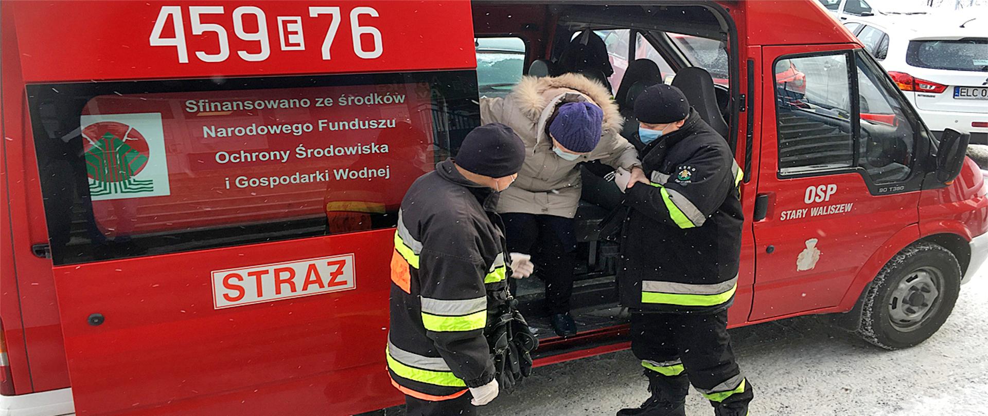 Na zdjęciu widać dwóch ratowników OSP pomagających wysiąść z auta starszej osobie. Jeden ratownik trzyma kobietę w podeszłym wieku za łokieć, drugi podaje jej rękę. Starsza osoba jest ubrana w zimową kurtkę jasnej barwy, czarną spódnicę, spodnie barwy czarnej oraz czapkę koloru fioletowego, na twarzy ma maseczkę jednorazową. Auto jest typu furgon barwy czerwonej z napisem na prawych przednich drzwiach OSP Stary Waliszew, po lewej stronie auta widnieje numer operacyjny samochodu oraz napis Straż. Ratownicy są ubrani w ubranie typu Nomex z odblaskami barwy jasnożółtej i szarej, na głowie mają czapkę wełnianą barwy czarnej, maseczkę na twarzy oraz rękawiczki jednorazowe. W tle samochody osobowe. Na ziemi widać śnieg.