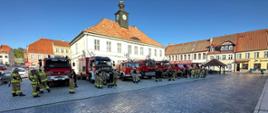Na placu stoi 9 samochodów ratowniczo-gaśniczych. Przed samochodami stoją strażacy. Przed strażakami droga brukowa. Za samochodami budynek ratusza w Reszlu.